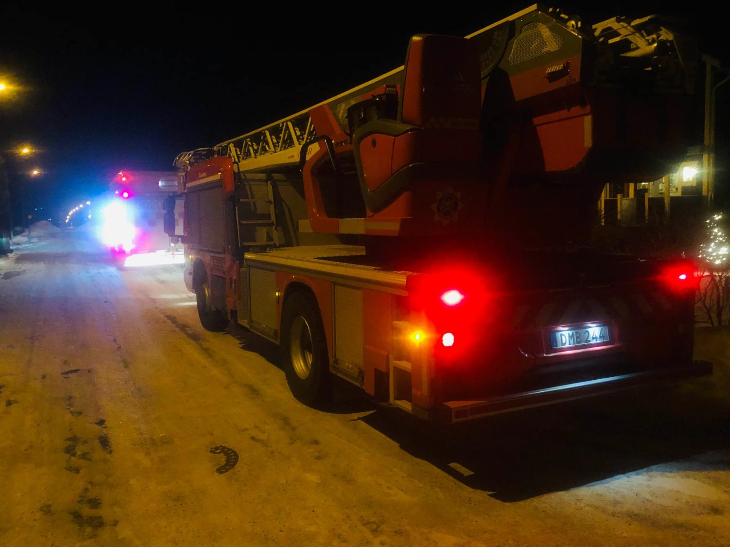 En brandbil med utskjuten stege står parkerad på en snöig gata på natten, med ett annat utryckningsfordon framför sig, båda med blinkande ljus.