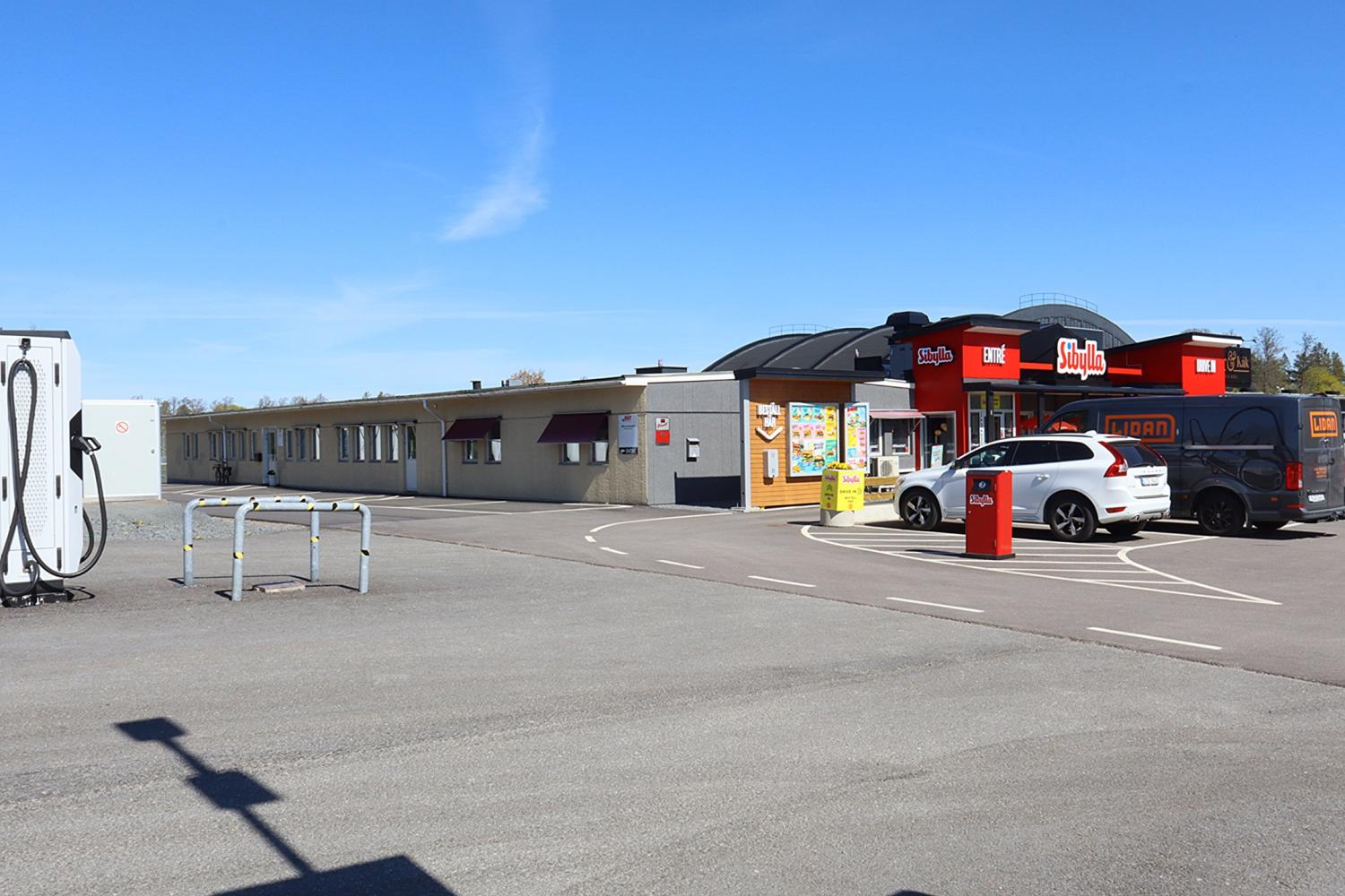 En bensinstation med närbutik och parkerade bilar en klar dag. Byggnaden har röda skyltar och stora fönster; en laddningsstation syns till vänster.