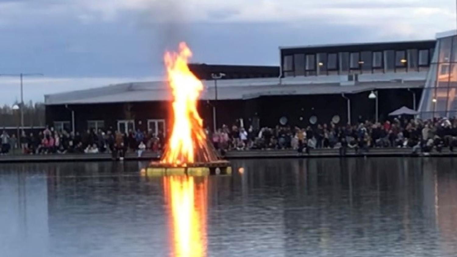 Brasa på en flytande plattform i en sjö med en folkmassa som tittar på från stranden i skymningen.