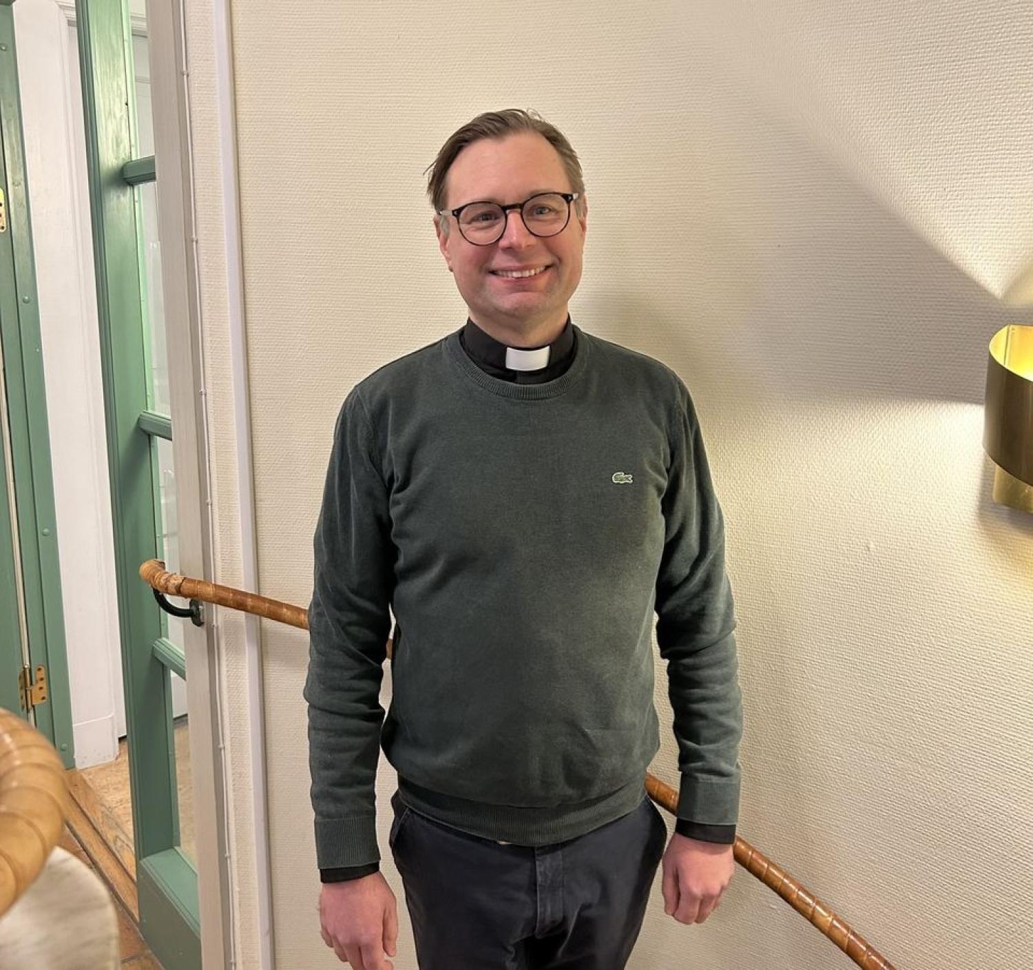 En man med glasögon och en grön tröja med kontorskrage står inomhus och ler mot kameran.
