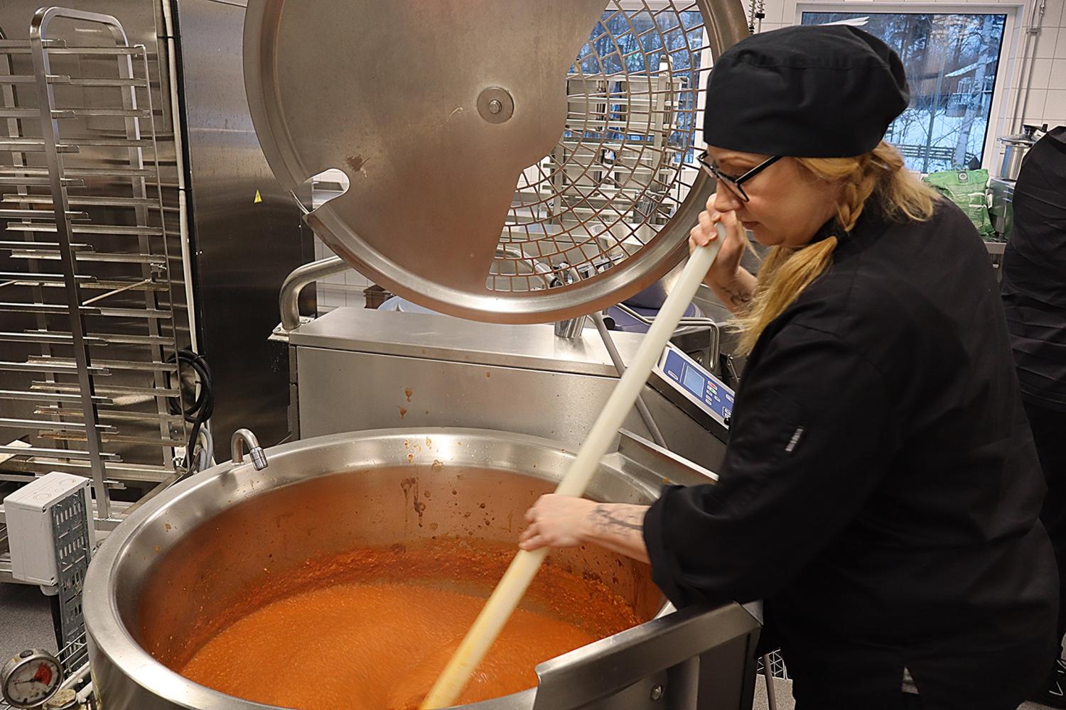 En kock i svart uniform och hatt rör om i en stor industriell gryta med tomatsås i ett storkök.