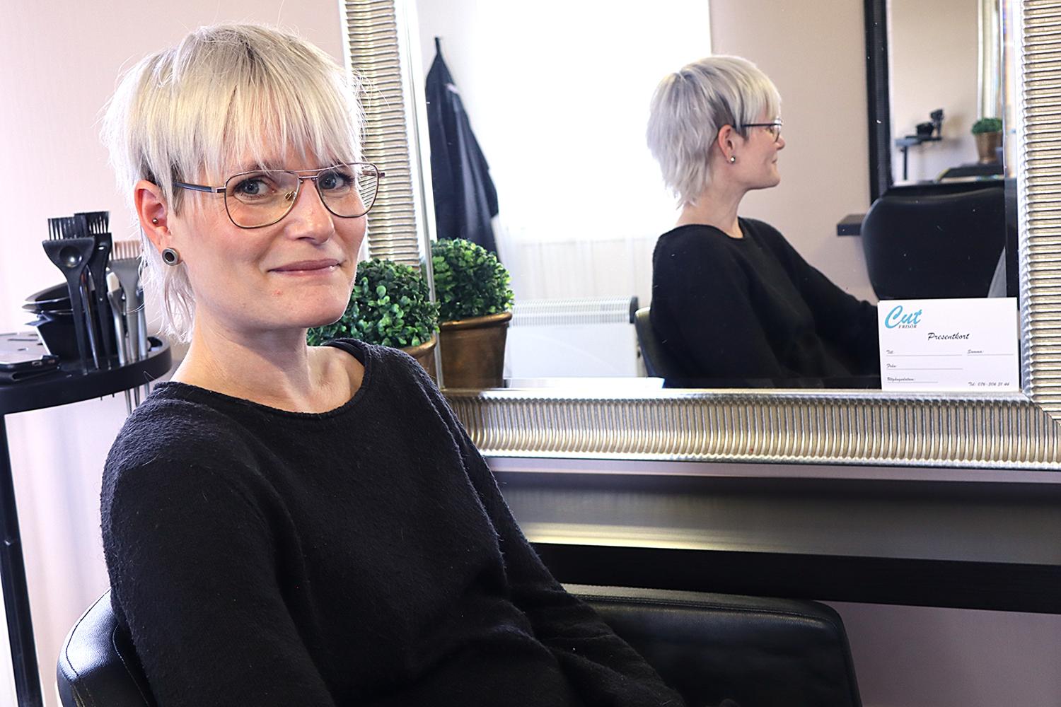 En kvinna med kort blont hår och glasögon sitter i en salongsstol vänd mot kameran; hennes spegelbild syns i spegeln bakom henne.