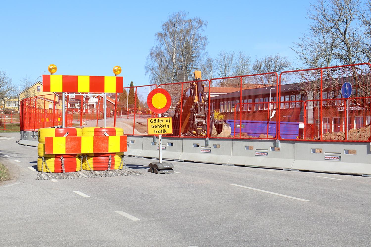 En väg är avstängd med röda och gula bommar, en skylt som begränsar obehörig trafik och byggutrustning som syns bakom stängslet en klar dag.