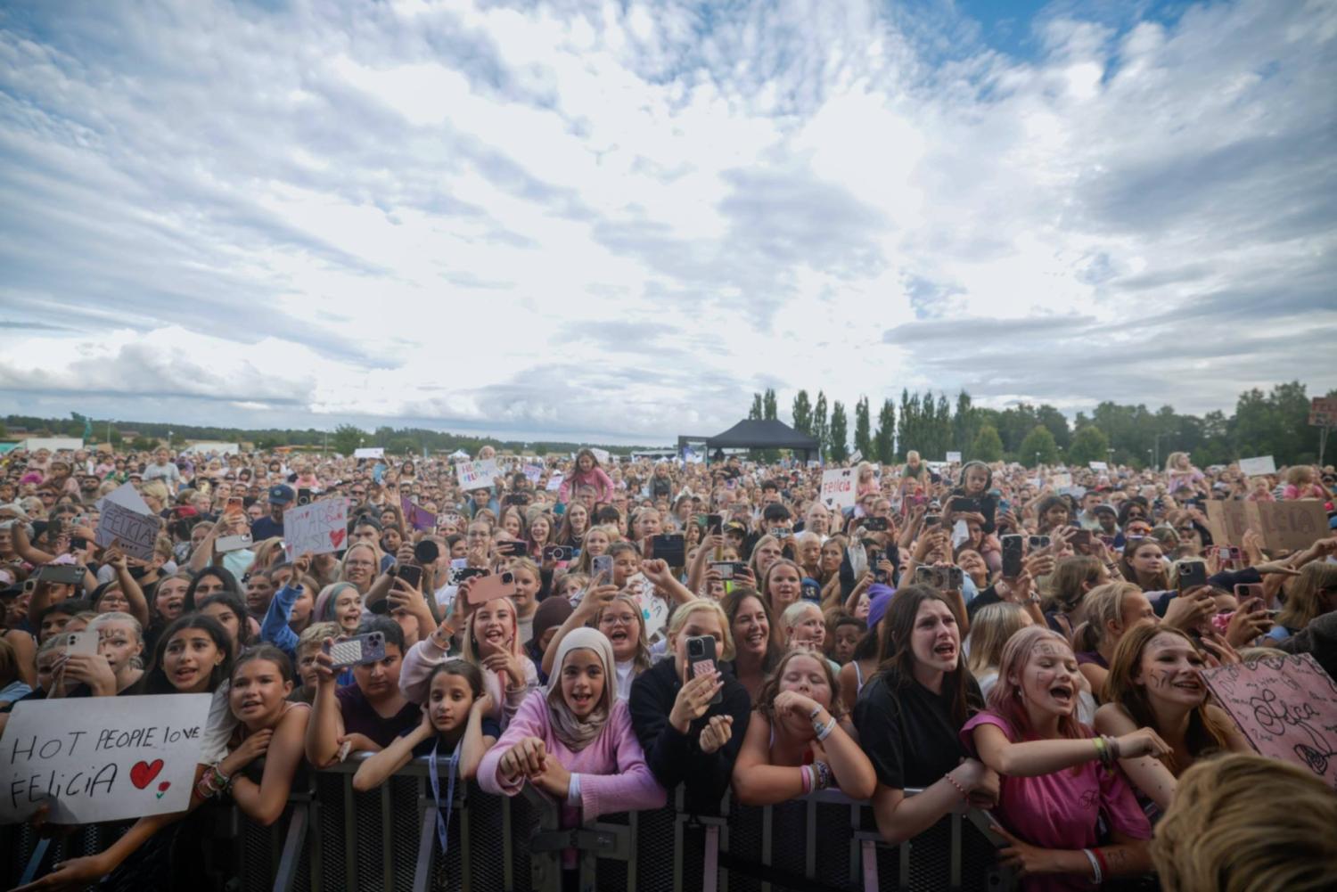 En stor skara entusiastiska fans, varav många håller upp skyltar och mobiltelefoner, står bakom en avspärrning vid en utomhuskonsert under en molnig himmel.