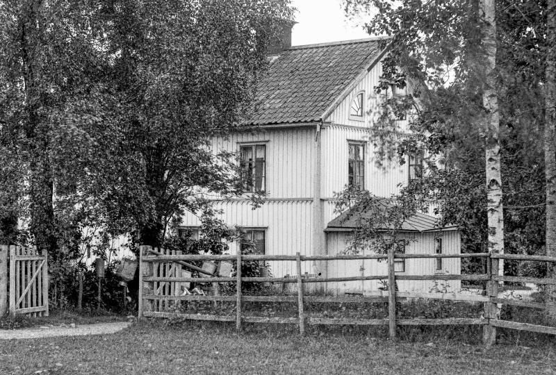 Svartvitt foto av ett tvåvåningshus i trä omgivet av träd och ett trästaket, med en grind synlig på vänster sida.