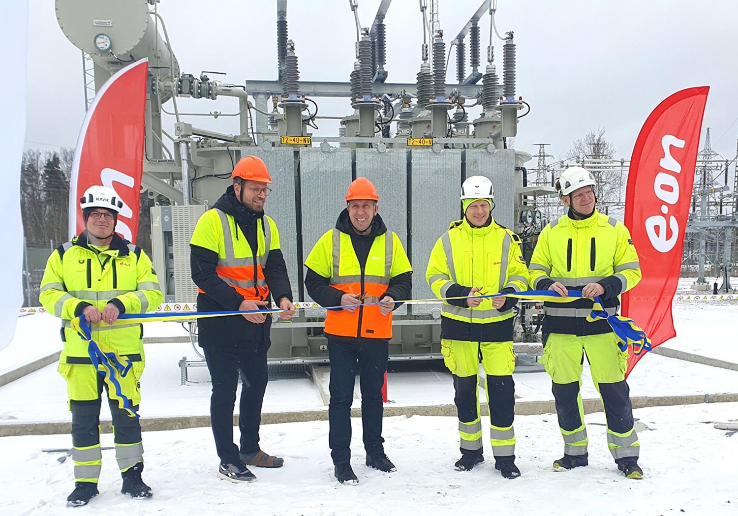 Fem personer i skyddsutrustning står ute i snön och håller i ett band vid en industrianläggning, med E.ON-banderoller och elektrisk utrustning i bakgrunden.
