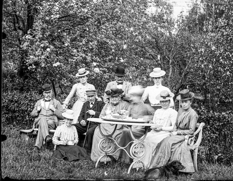 En grupp män och kvinnor i vintagekläder sitter och står runt ett trädgårdsbord utomhus, med träd och buskar i bakgrunden och en hund som ligger i förgrunden.
