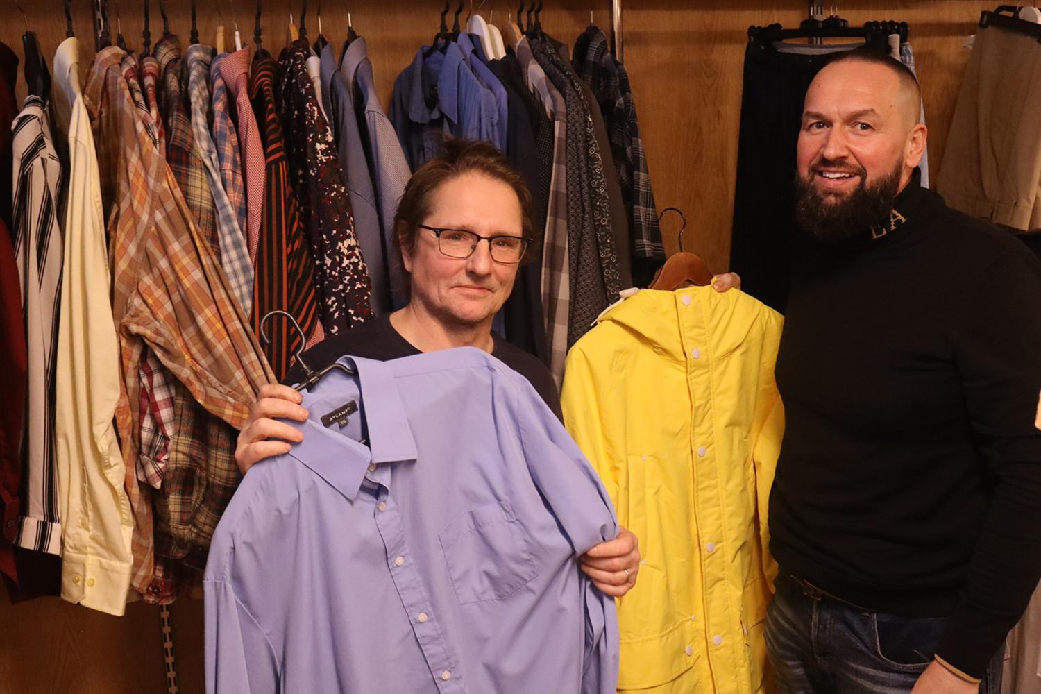Två män står framför en garderob med kläder; den ene håller i en blå skjorta, den andre håller i en gul regnrock.