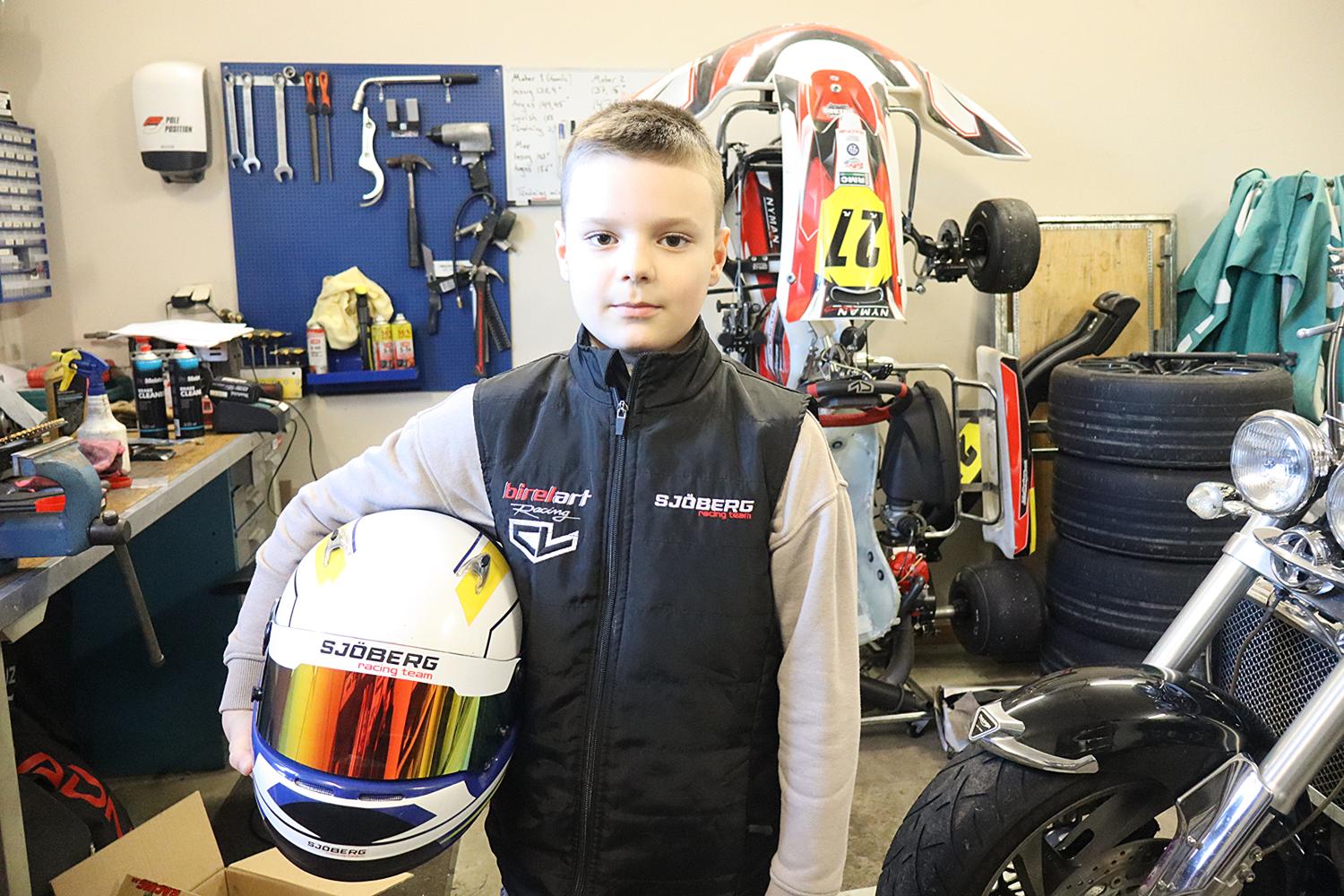 En pojke står i en verkstad och håller i en racinghjälm, med verktyg, däck och en gokart i bakgrunden.