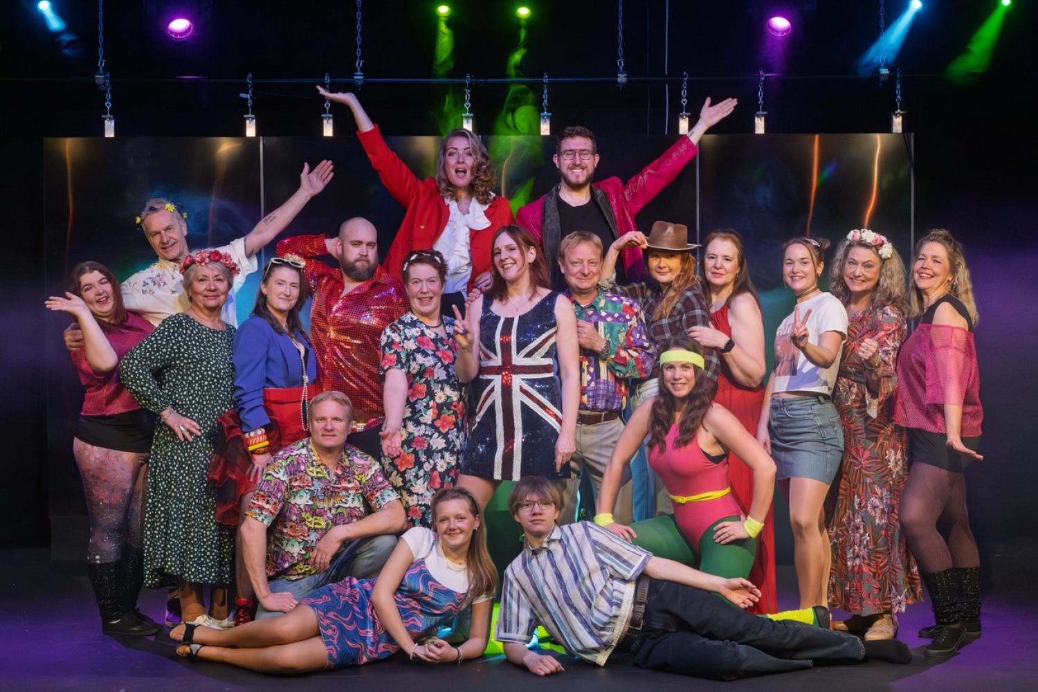 En grupp människor i färgglada retrokostymer poserar tillsammans på scenen i starkt ljus, några står, sitter och höjer armarna, framför en mörk bakgrund.