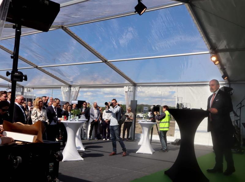 En man i kostym talar på ett podium under ett genomskinligt tält till en publik; fotografer och en kameraman fångar händelsen.