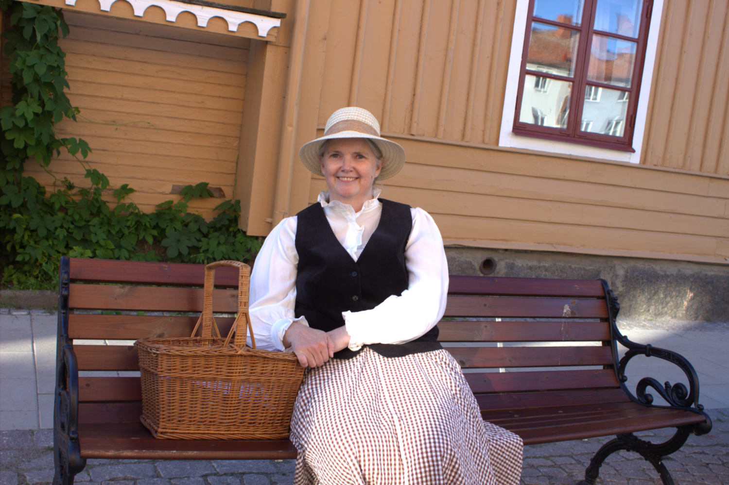 En kvinna i traditionella kläder och med en stråhatt sitter på en träbänk bredvid en flätad korg, med en träbyggnad i bakgrunden.