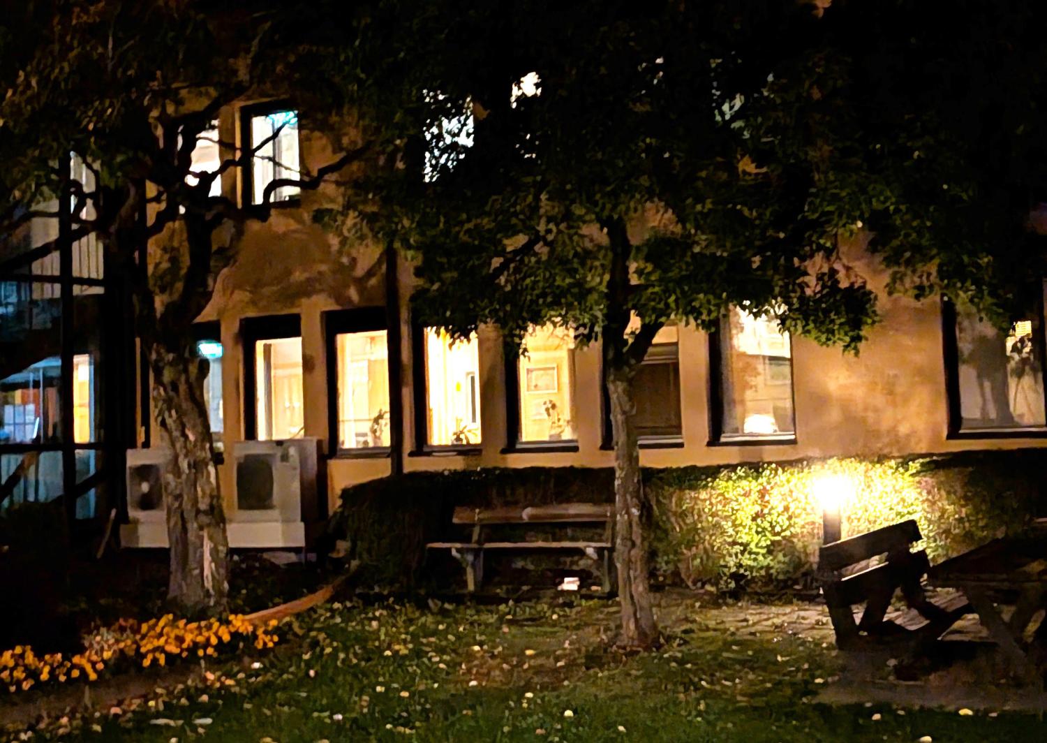En svagt upplyst trädgård med bänkar och träd utanför en byggnad vars fönster lyser upp på natten.