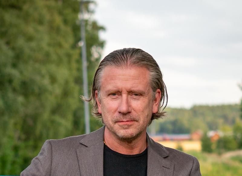 En medelålders man med bakkammat hår och ett välansat skägg bär en grå kavaj och en svart skjorta och står utomhus med träd och byggnader i bakgrunden.