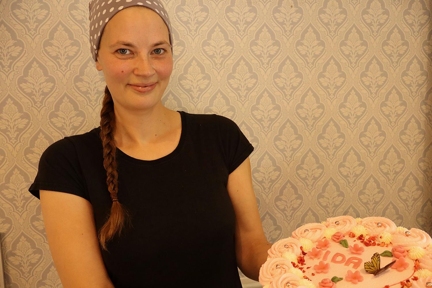 En kvinna med prickig huvudduk och svart skjorta håller en rosa frostad tårta dekorerad med namnet 