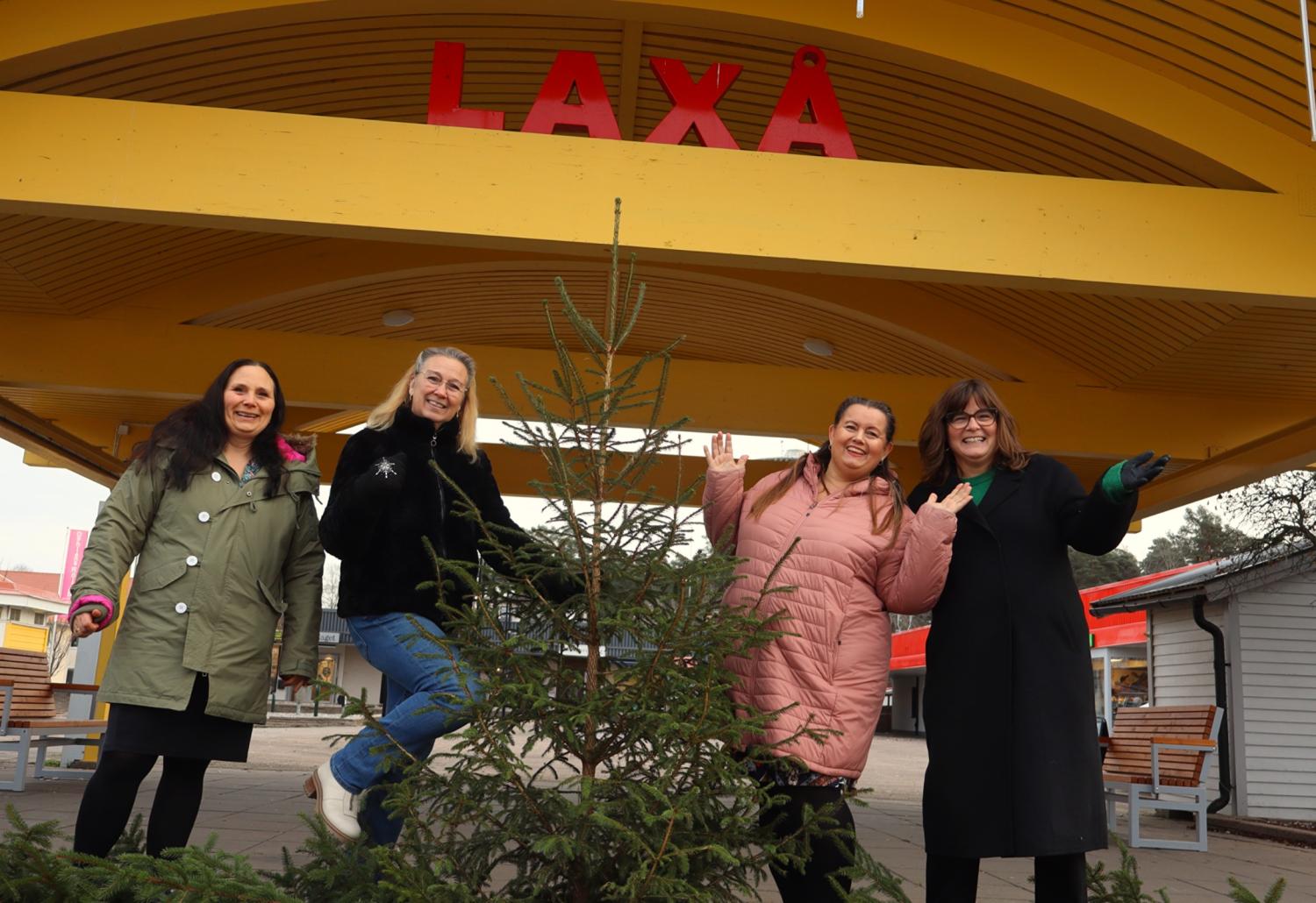 Fyra kvinnor står och poserar runt en liten julgran under en gul paviljong med ordet 