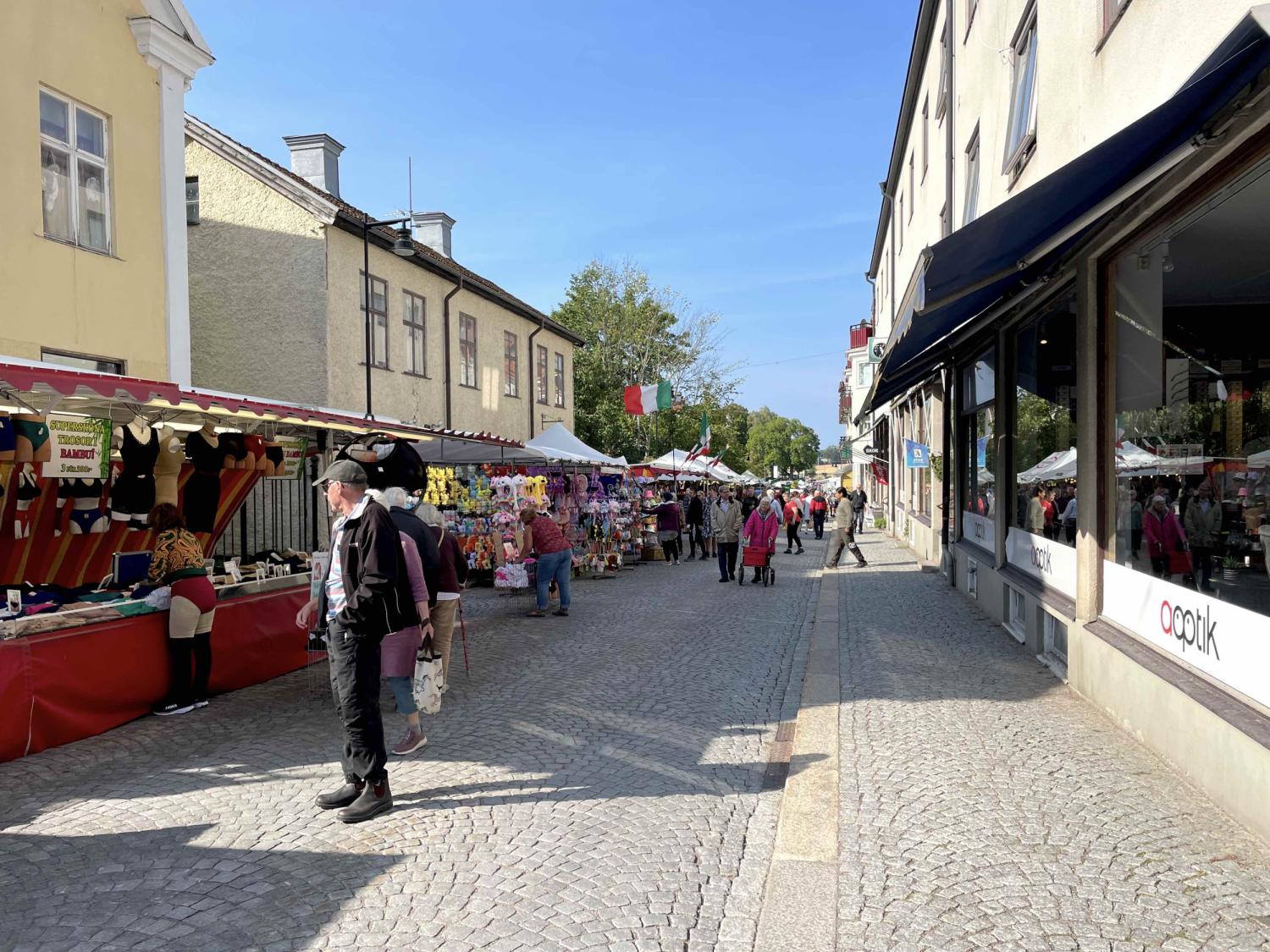 En gatumarknad med olika stånd som säljer varor, människor som går och shoppar, byggnader längs båda sidorna och en solig himmel ovanför.