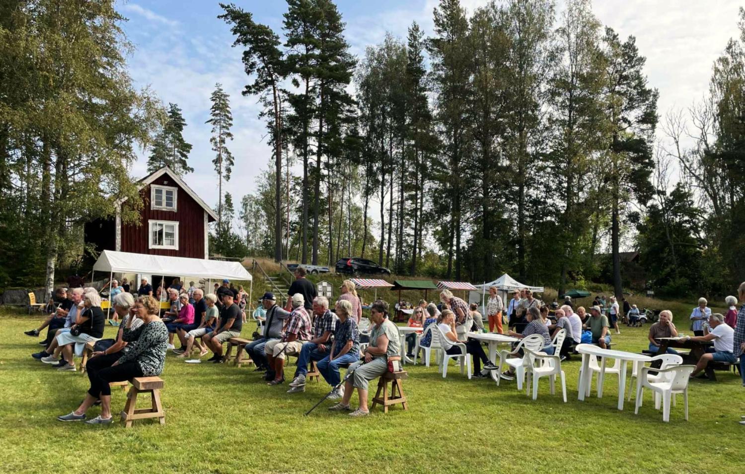 En grupp människor sitter på stolar och bänkar utomhus på en gräsplan nära ett rött hus och några tält, med träd i bakgrunden.