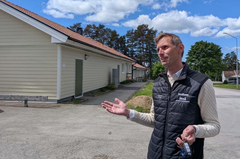 En man i svart väst står utanför ljusa byggnader en solig dag och gestikulerar med händerna samtidigt som han håller i en nyckelknippa.