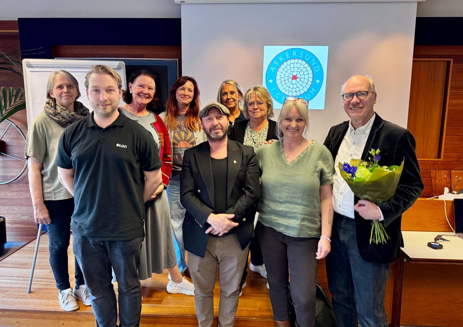 En grupp på tio personer står inomhus framför en presentationsskärm och en person håller i en blombukett.
