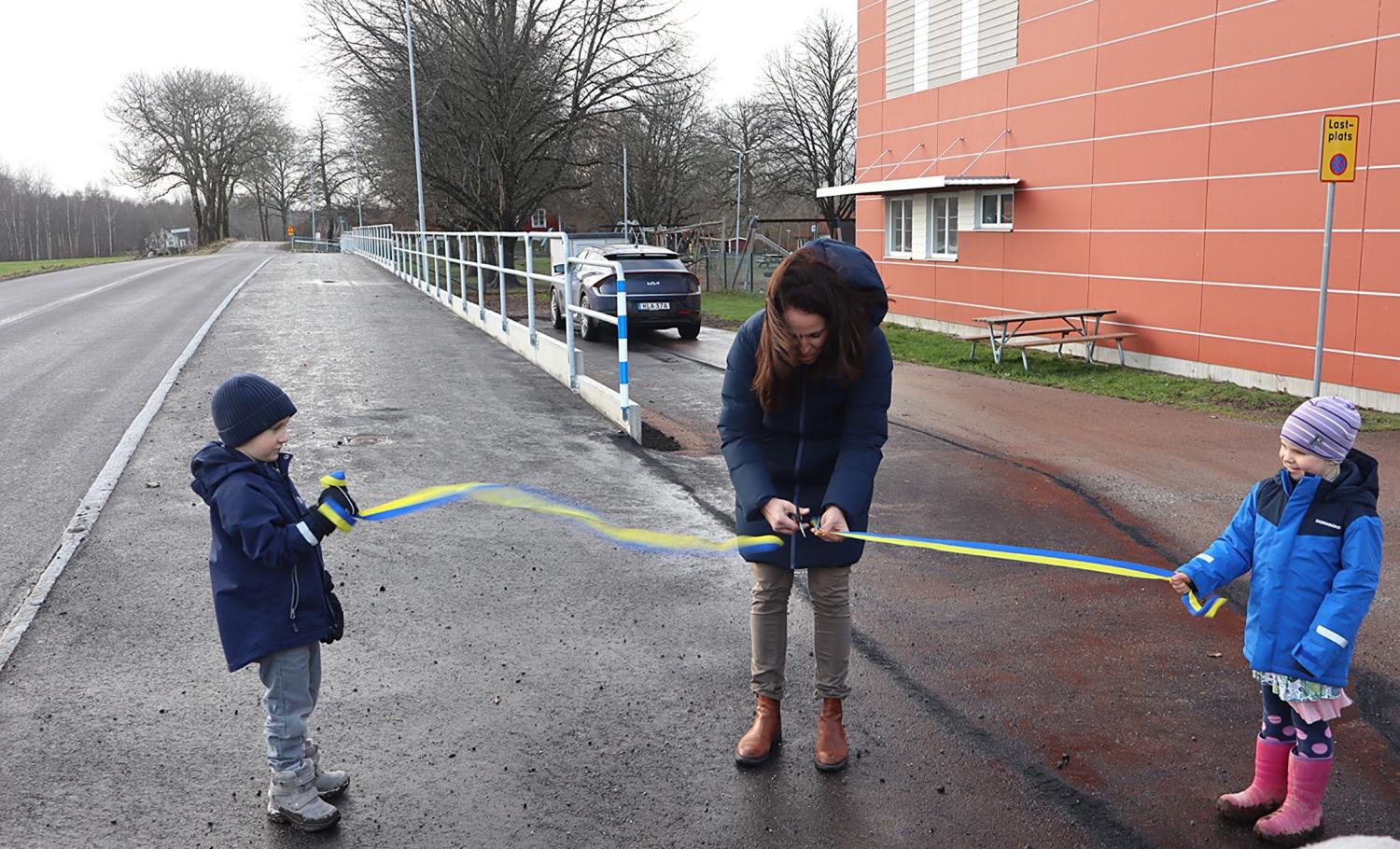 En kvinna klipper av ett blågult band som hålls av två barn vid en vägkant intill en byggnad för att markera en invigning.