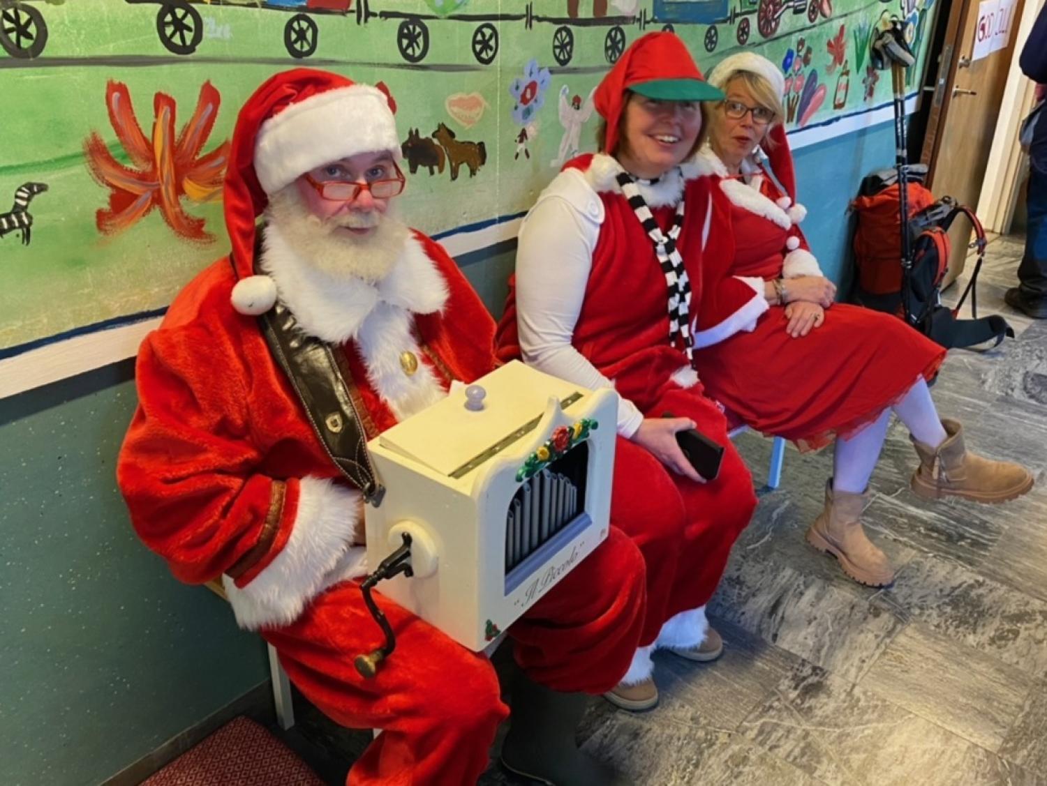 Tre personer i festliga dräkter, varav en är utklädd till tomte och håller i en vit låda, sitter på en bänk i en dekorerad hall med en väggmålning bakom sig.