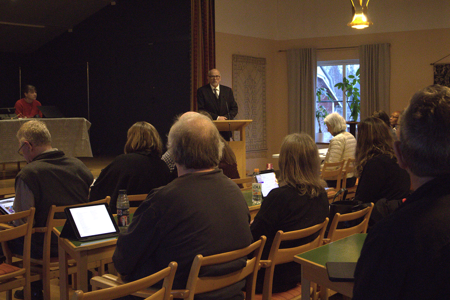 En man står på ett podium och talar till en publik som sitter vid bord med bärbara datorer och anteckningsböcker i ett konferensrum.
