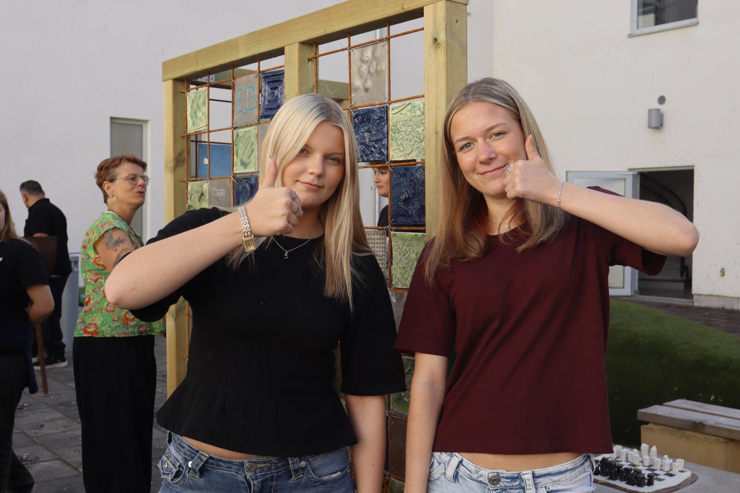 Två unga kvinnor står utomhus framför en kakelutställning och gör båda tummen upp-tecknet med höger hand nära hakan.