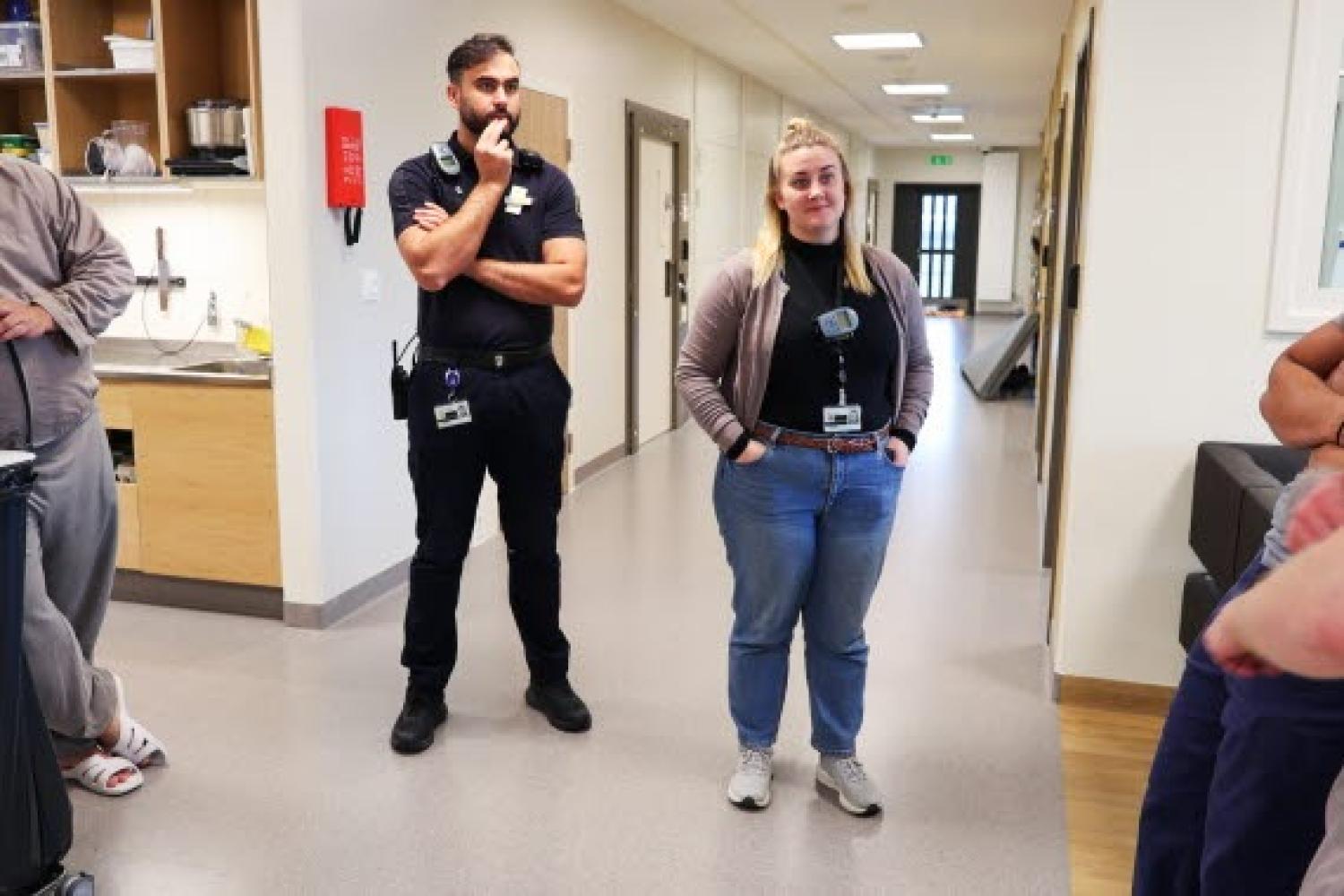 Två personer med arbetsmärken står i en korridor, den ena med armarna i kors och den andra med händerna i fickorna. Andra personer är delvis synliga i närheten.