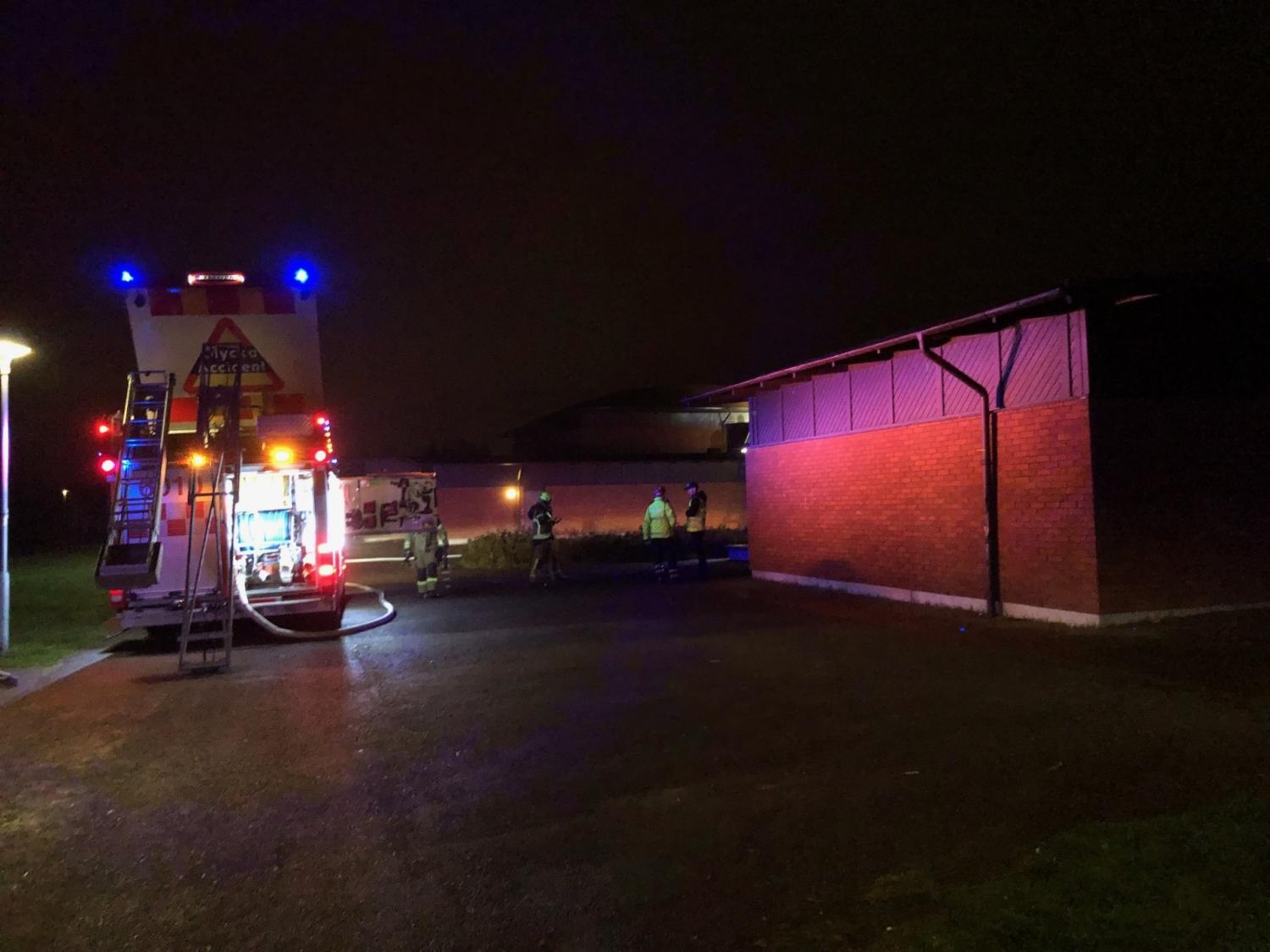 En brandbil med tända lampor står parkerad utanför en tegelbyggnad på natten, med brandmän och slangar synliga i närheten av platsen.