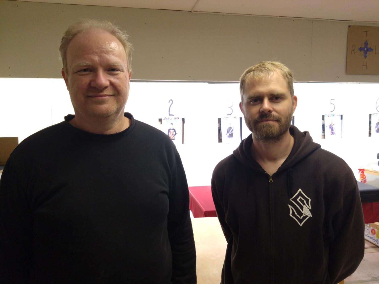 Två män som står inomhus framför en vägg med uppsatta pappersark. Mannen till vänster har en svart skjorta, medan mannen till höger har en svart luvtröja.