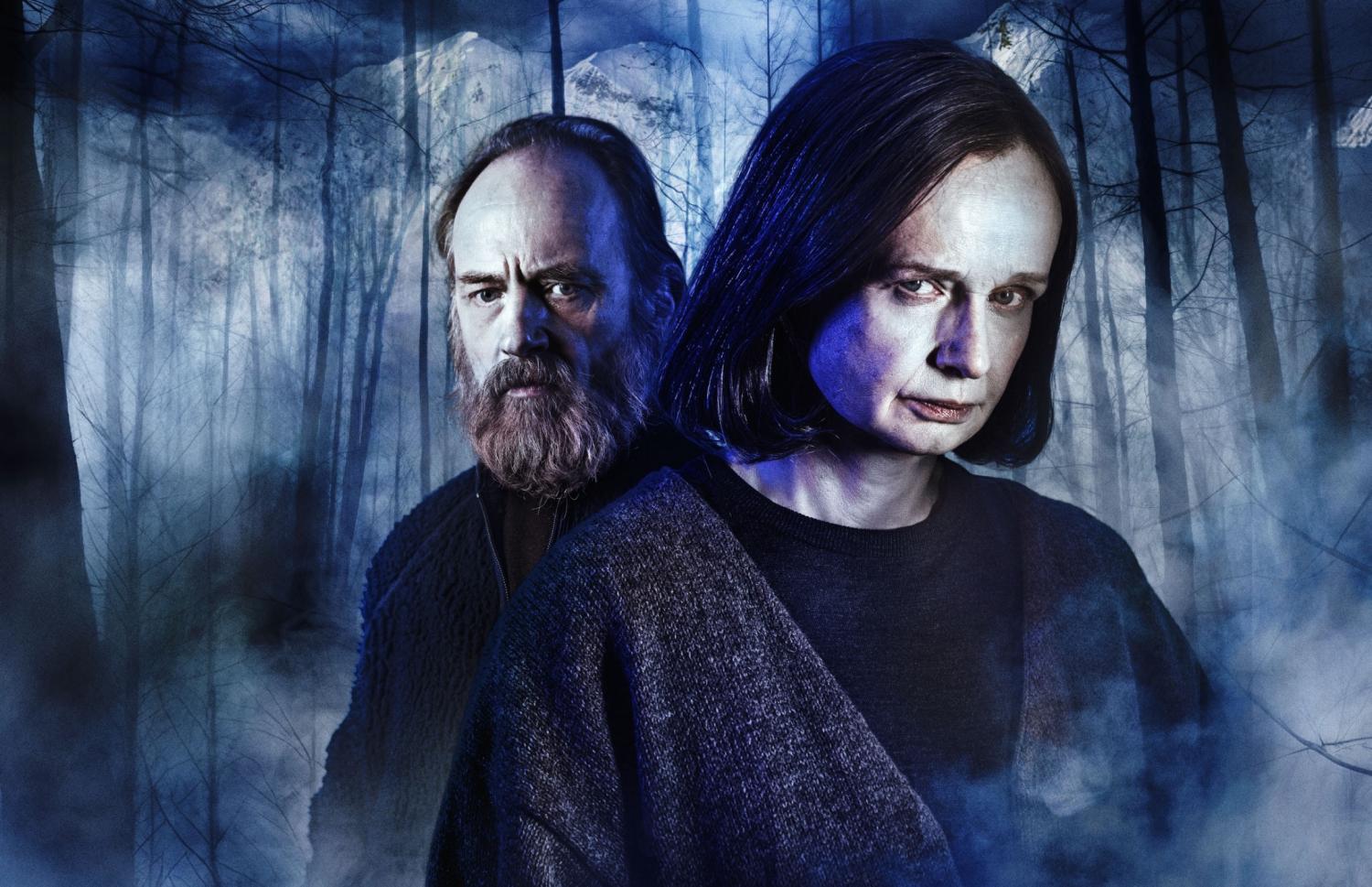 Två allvarliga personer i mörka kläder står rygg mot rygg i en dimmig, blåbelyst skog med kala träd och en bergig bakgrund.