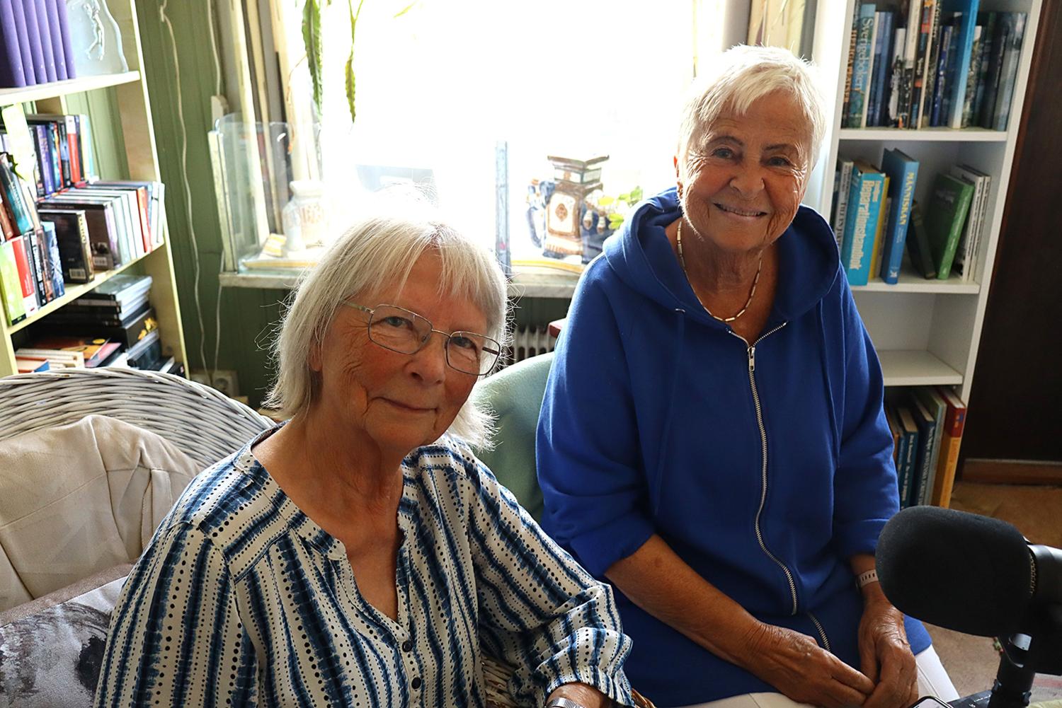 Två äldre kvinnor sitter inomhus i ett mysigt rum med bokhyllor och ler mot kameran. Den ena bär en randig skjorta, den andra en blå zip-up topp. En mikrofon står i förgrunden.
