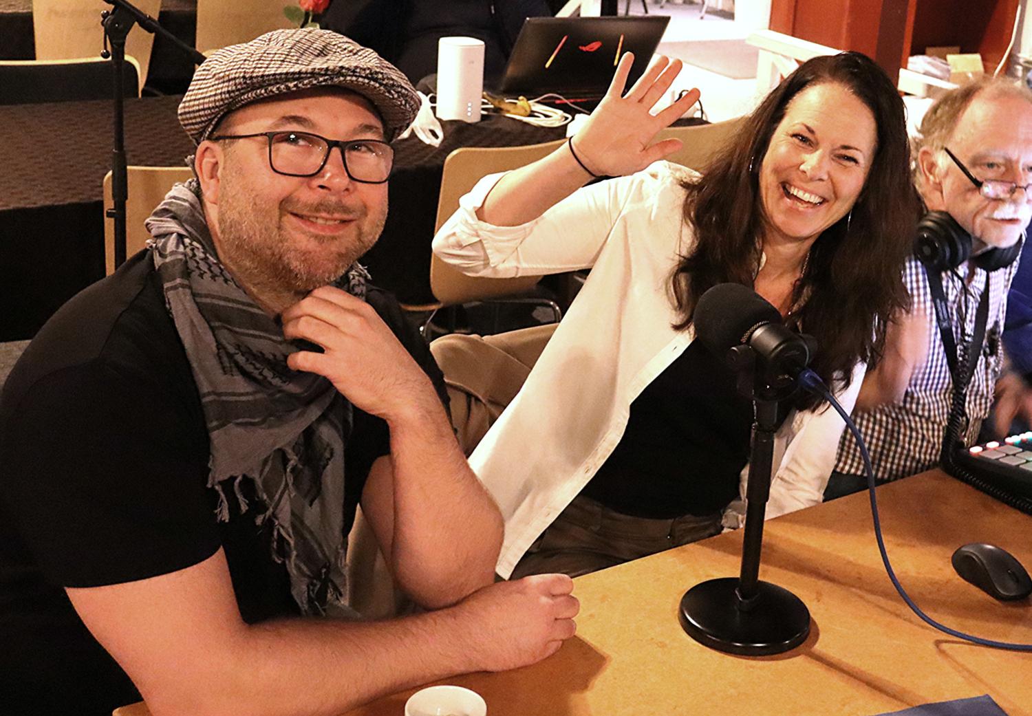 Tre vuxna sitter vid ett bord med mikrofoner och ljudutrustning; en kvinna vinkar och ler, medan de andra tittar mot kameran.