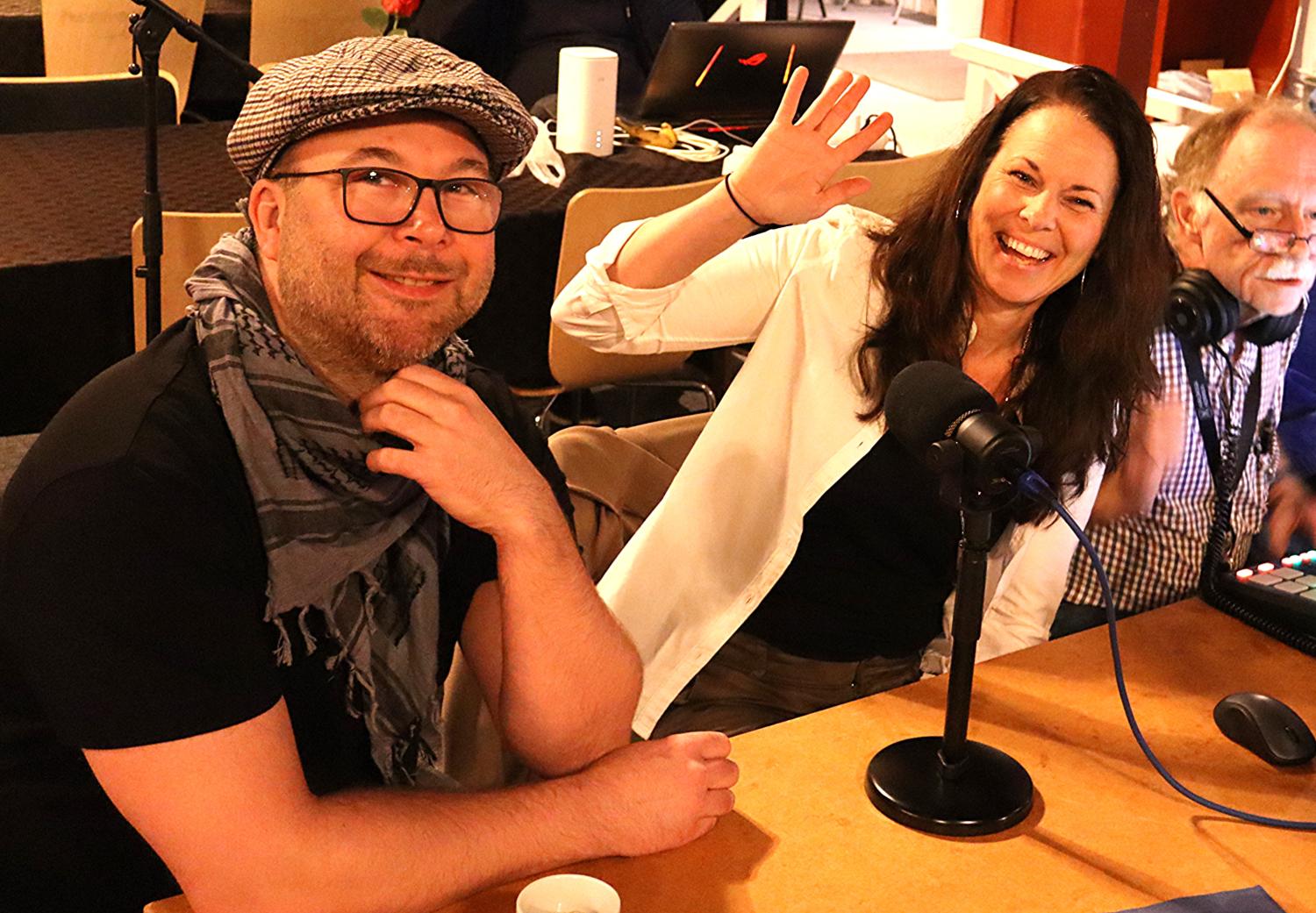 Tre vuxna sitter vid ett bord med mikrofoner och ljudutrustning; en kvinna vinkar och ler mot kameran medan de andra tittar på.