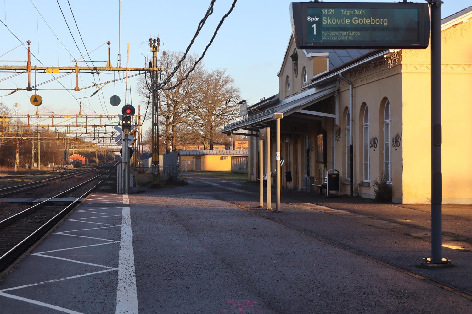Tom tågstationsplattform med en skylt som visar tåginformation för Skövde och Göteborg; spår och kontaktledningar synliga under en klar dag.