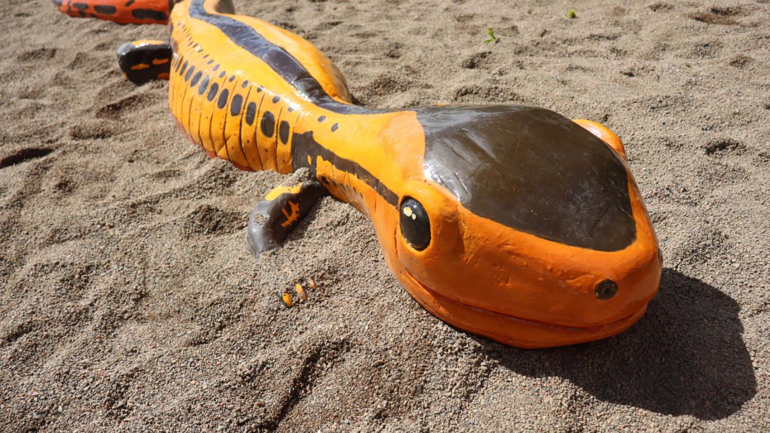 Stor skulptur föreställande en orange och svart salamander på sandig mark, sedd från sidan, med synliga fläckar och detaljer på kroppen.