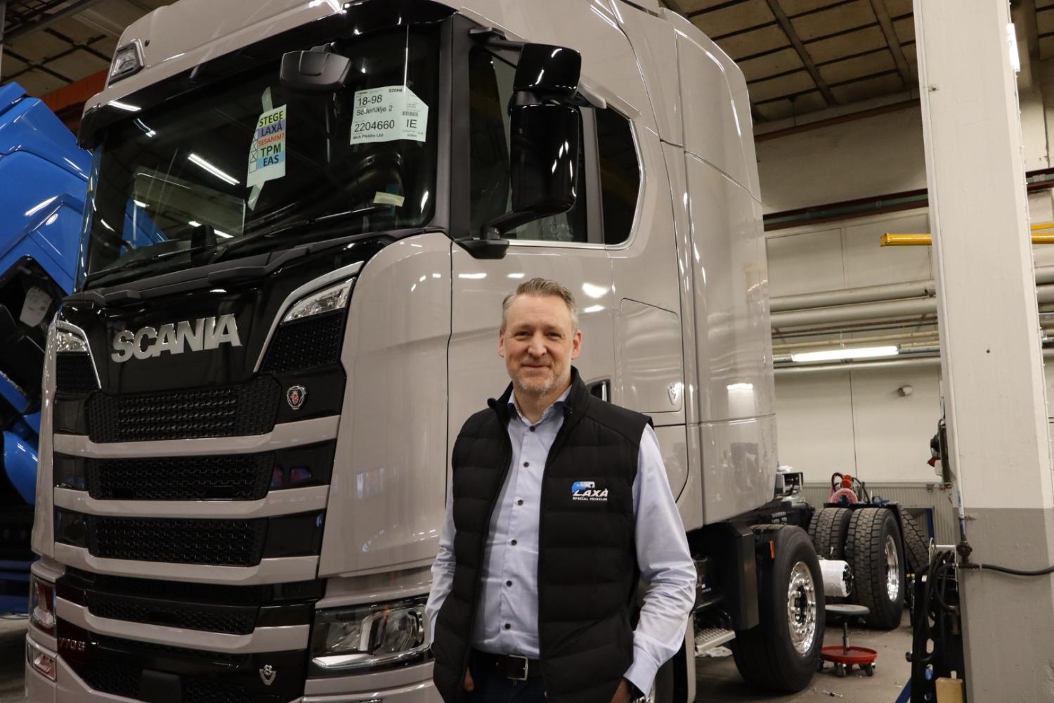 En person som står framför en stor grå Scania-lastbil inne i ett lager, iklädd svart väst och blå skjorta.