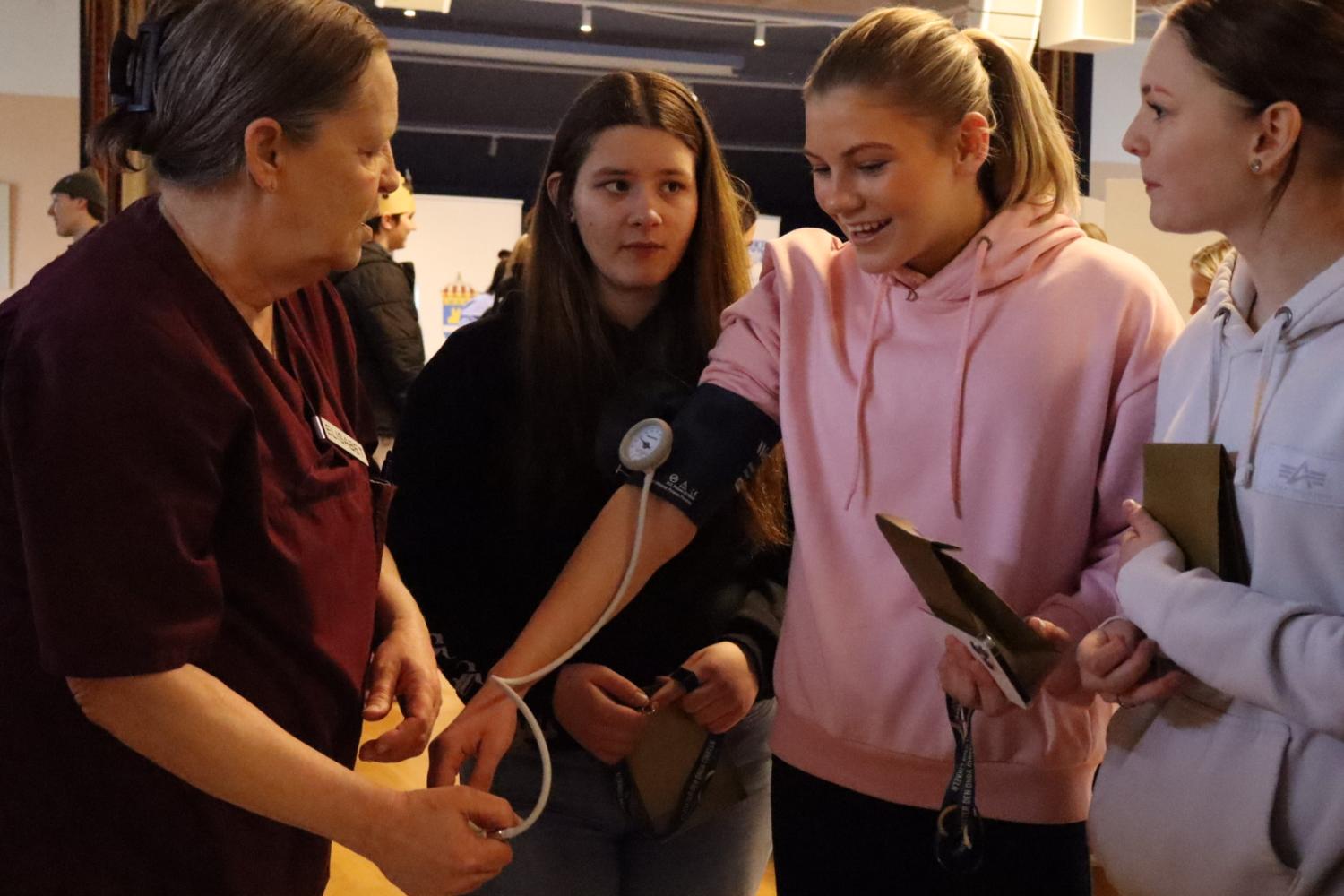 En kvinna i medicinsk uniform hjälper en ung kvinna med en blodtrycksmätare, medan två andra unga kvinnor observerar.