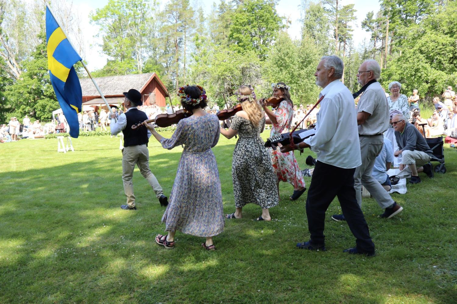 En grupp människor i traditionella kläder spelar fioler och går på gräs, ledda av en person som bär en svensk flagga, med en publik sittande i bakgrunden.