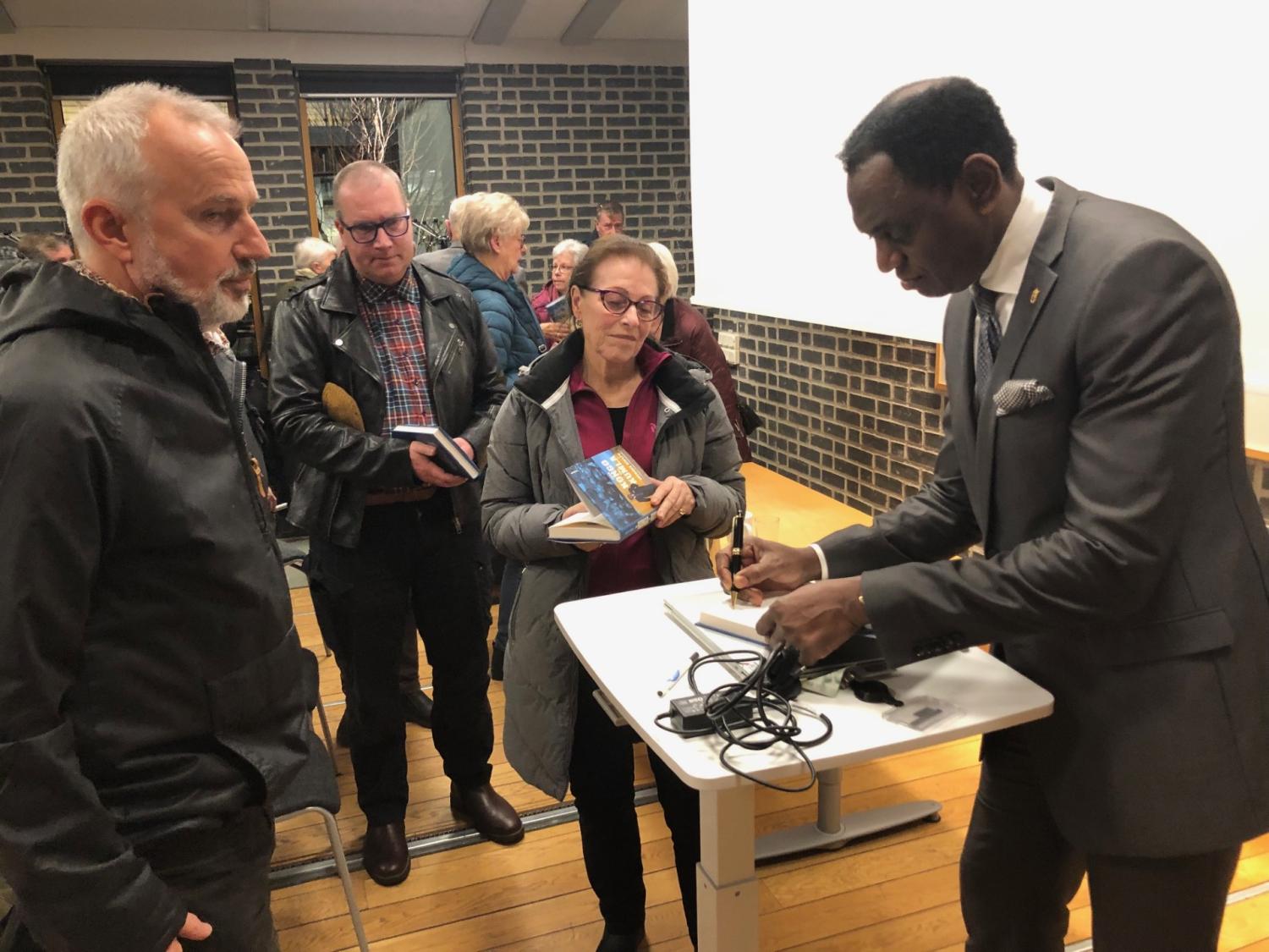 En man i kostym signerar en bok vid ett bord, medan tre personer står i kö och håller i böcker.