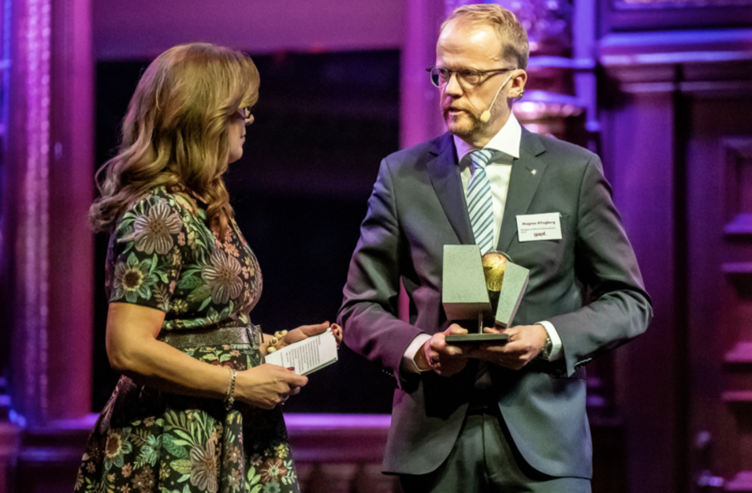En man och en kvinna i högtidlig klädsel står på en scen. Mannen håller en liten utmärkelse.