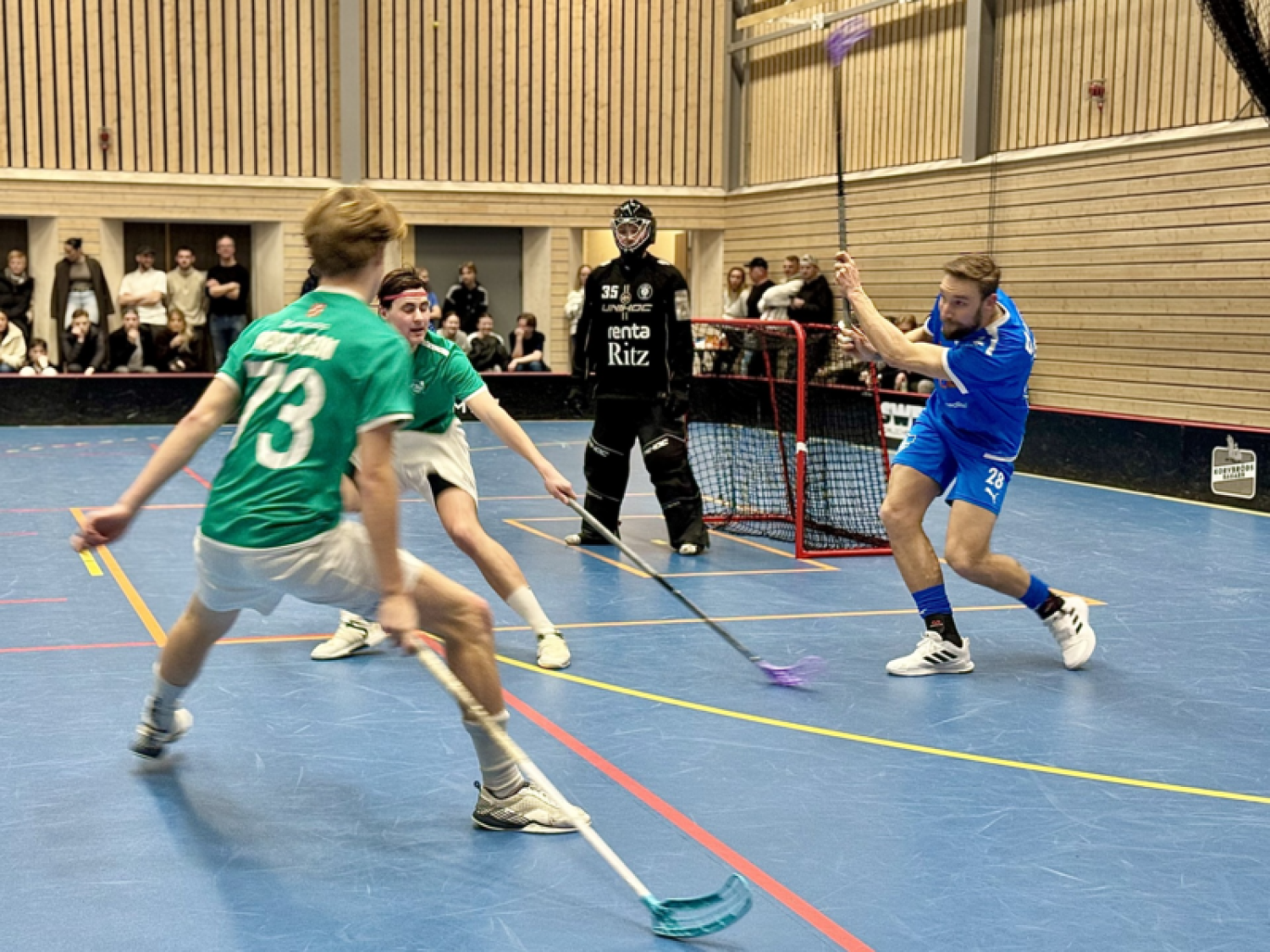 Två innebandyspelare i gröna tröjor försvarar sig mot en spelare i blå tröja på väg att skjuta. Målvakten i svart utrustning står redo framför nätet. Åskådare tittar i bakgrunden.