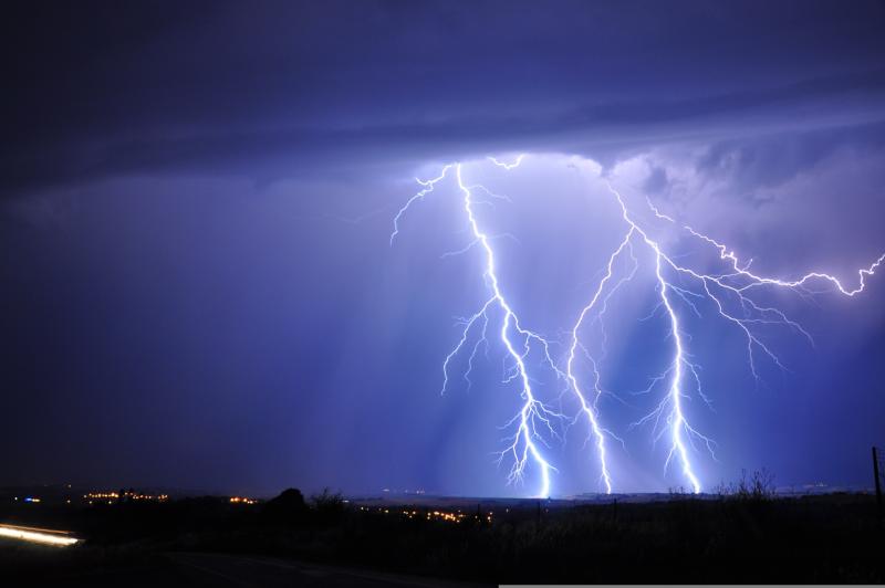 Flera blixtar slår ner i marken under mörka stormmoln på natten, och stadens ljus syns vid horisonten.