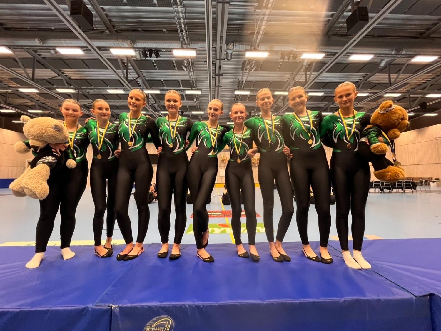 Nio kvinnliga gymnaster i matchande gröna och svarta trikåer står på ett podium med medaljer och uppstoppade djur i en gymnastiksal.