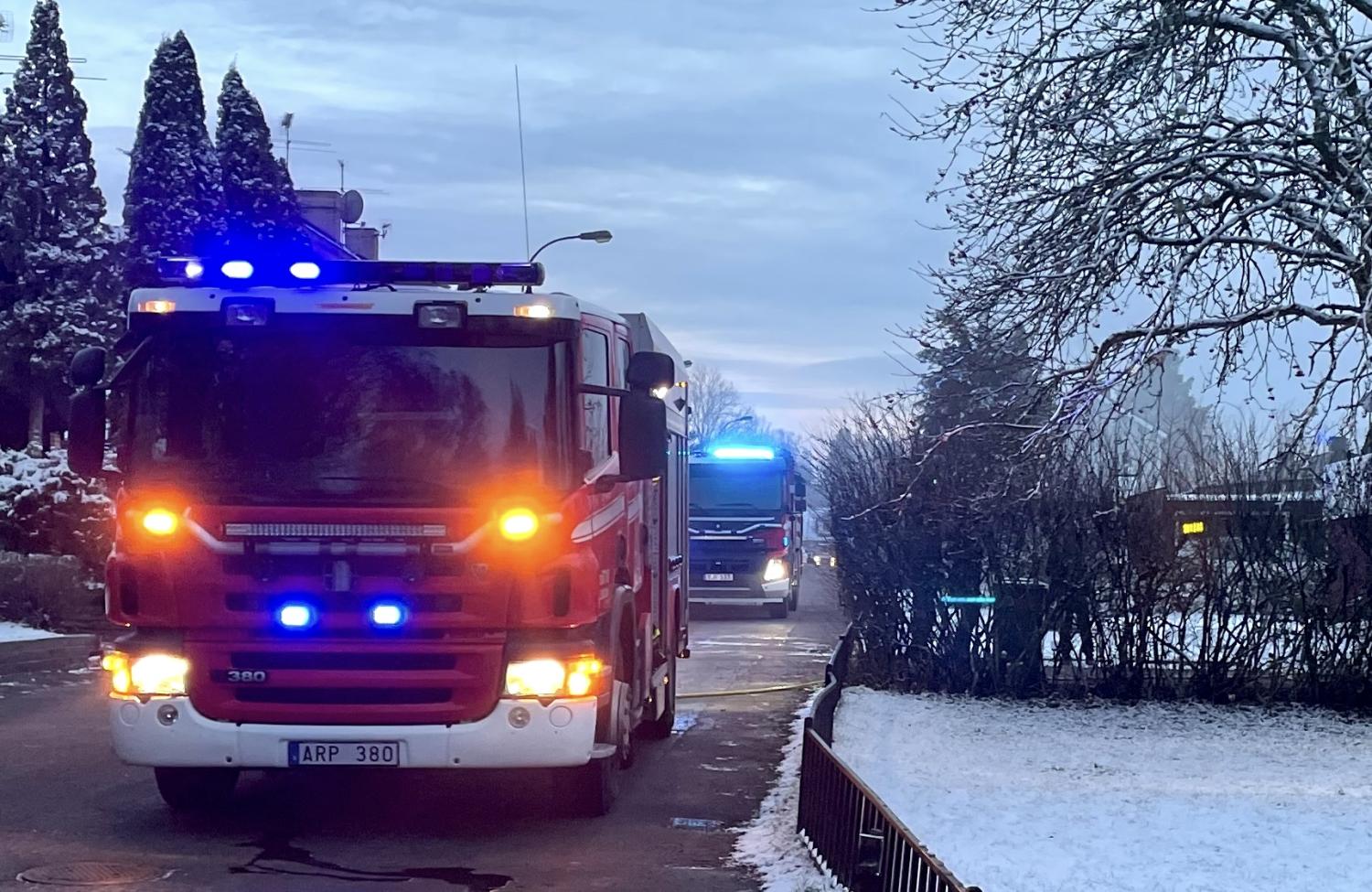 Brandbilar med blinkande ljus parkerade på en snöig gata kantad av träd och buskar.