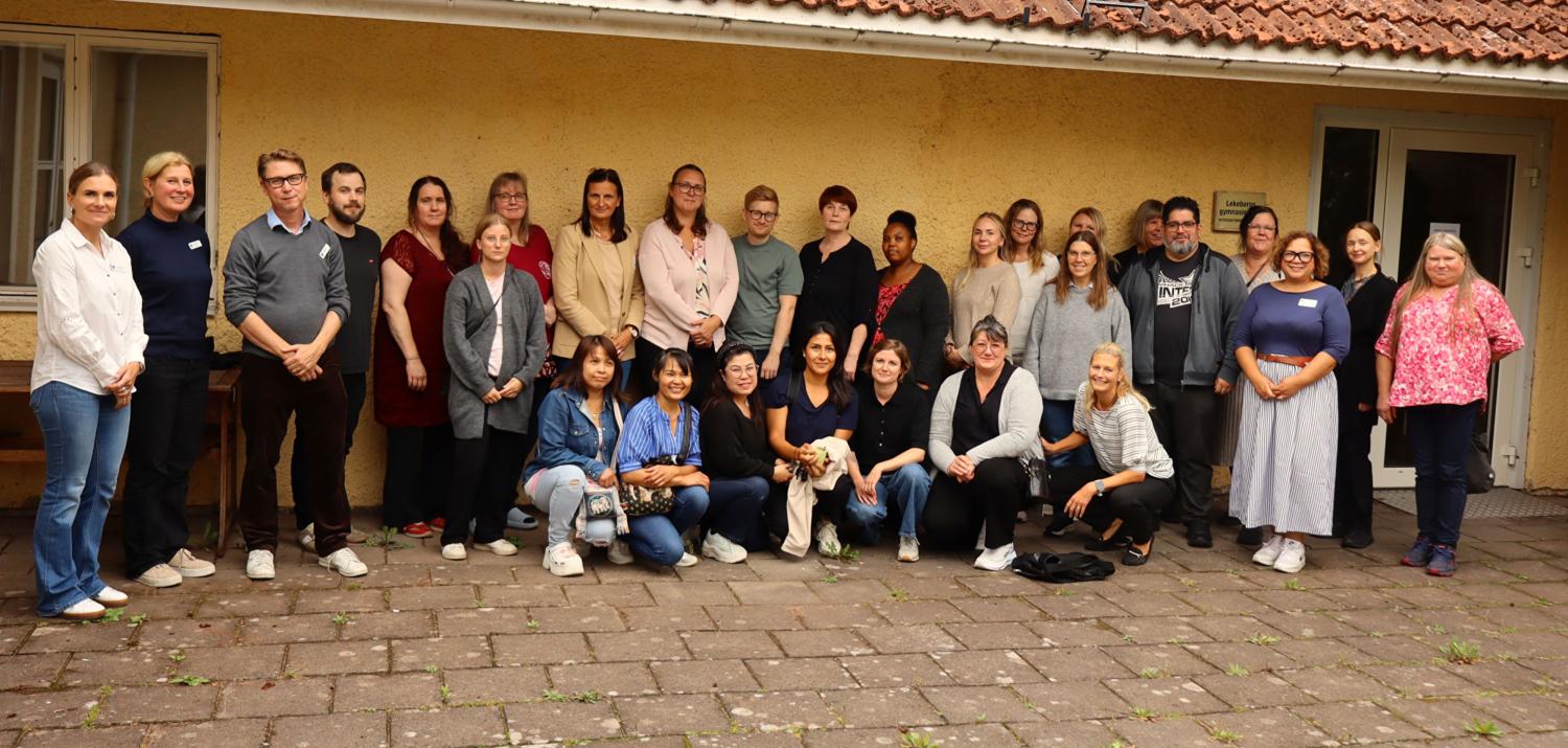 En grupp vuxna poserar tillsammans utomhus framför en gul byggnad, stående och sittande på en asfalterad yta.