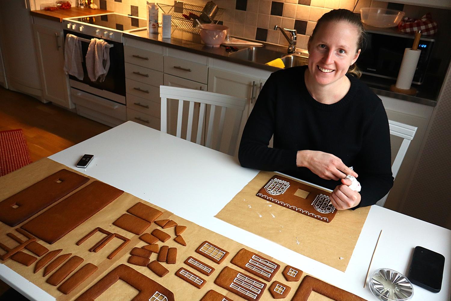 En kvinna sitter vid ett köksbord och sätter ihop delar till ett pepparkakshus, med glasyr i handen och olika dekorerade pepparkaksplattor framför sig.