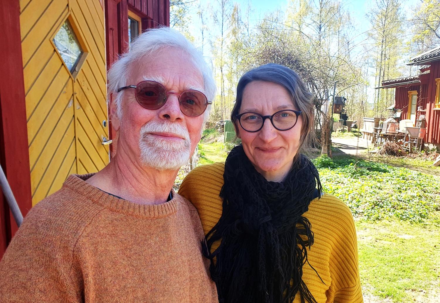 En äldre man och en kvinna står tätt intill varandra utomhus vid en röd träbyggnad och ler mot kameran en solig dag.