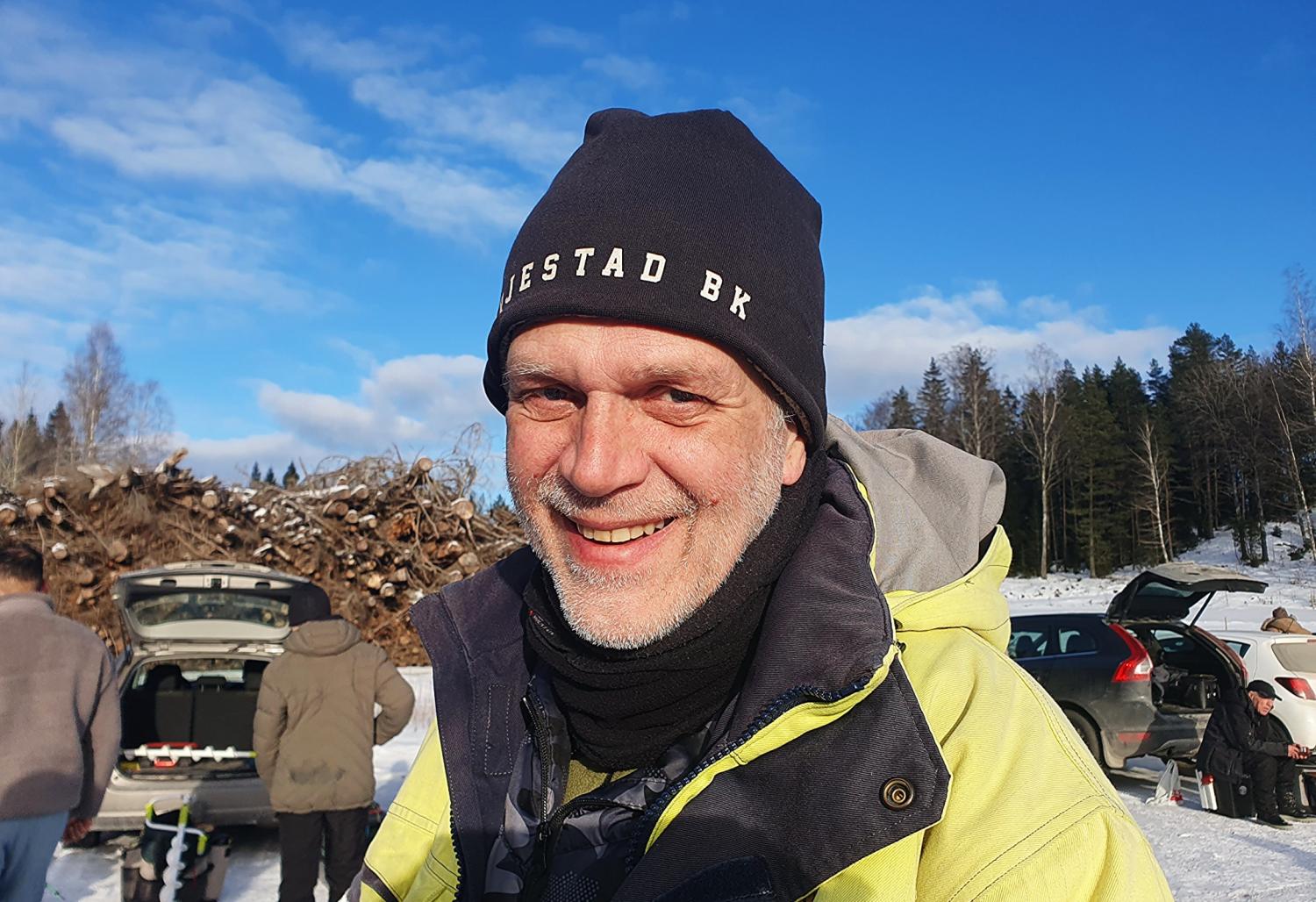 En man med svart mössa och gul jacka ler utomhus i ett snöigt område, med bilar och människor i bakgrunden under en blå himmel.